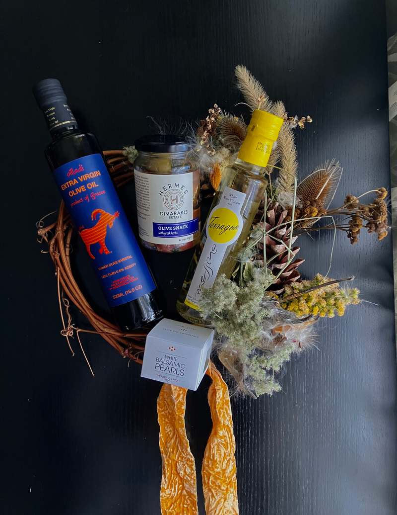 A collection of gourmet greek products including a bottle of extra virgin olive oil, a jar of olives, a bottle of white wine vinegar with Tarragon and balsamic pearls
