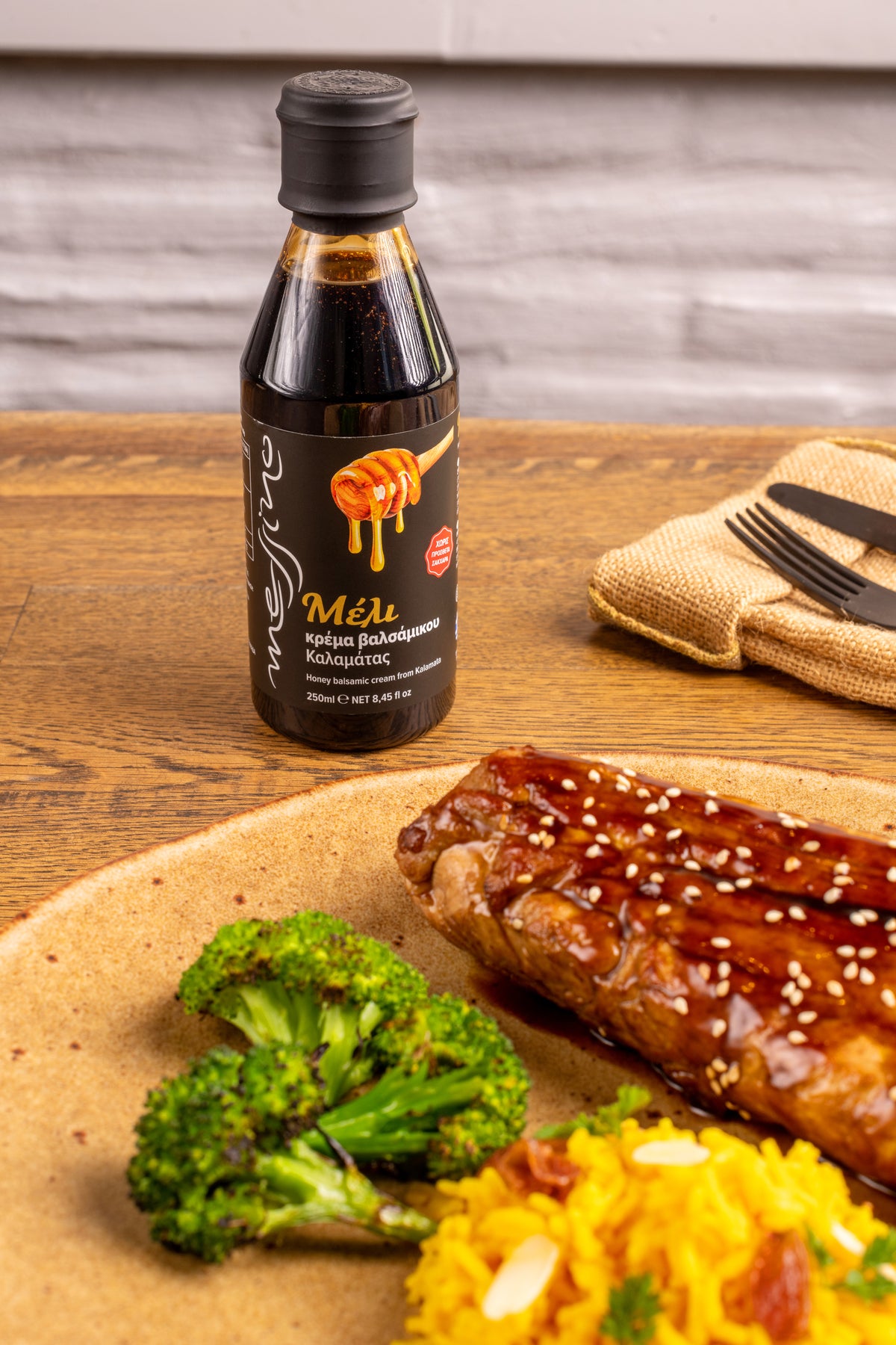 Bottle of Messino honey Balsamic Glaze on a wooden table with grilled salmon, broccoli, and yellow rice.